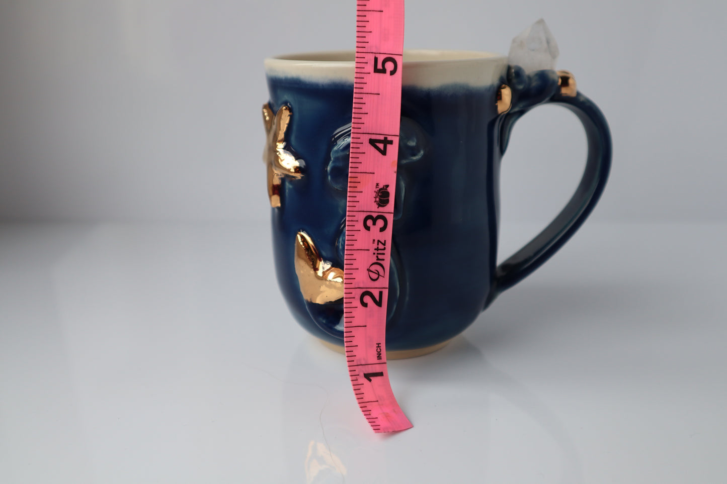 Blue mug with gold accents and a pink measuring tape for scale on a white background