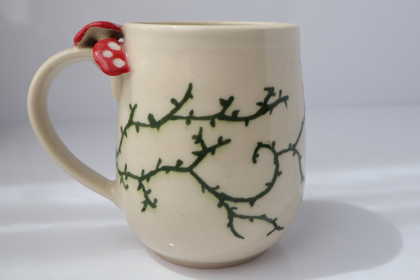 Beige mug with green branch design and red mushroom handle on a white background.