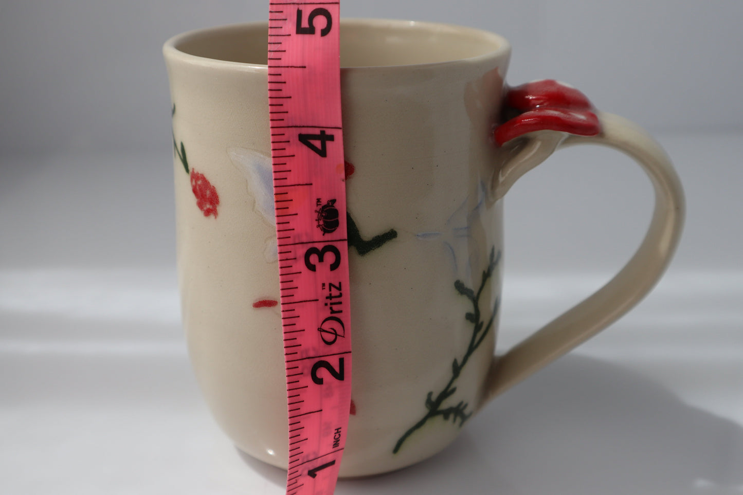 Ceramic mug with a fairy design and red mushroom handle, measured against a pink tape measure on a white background.