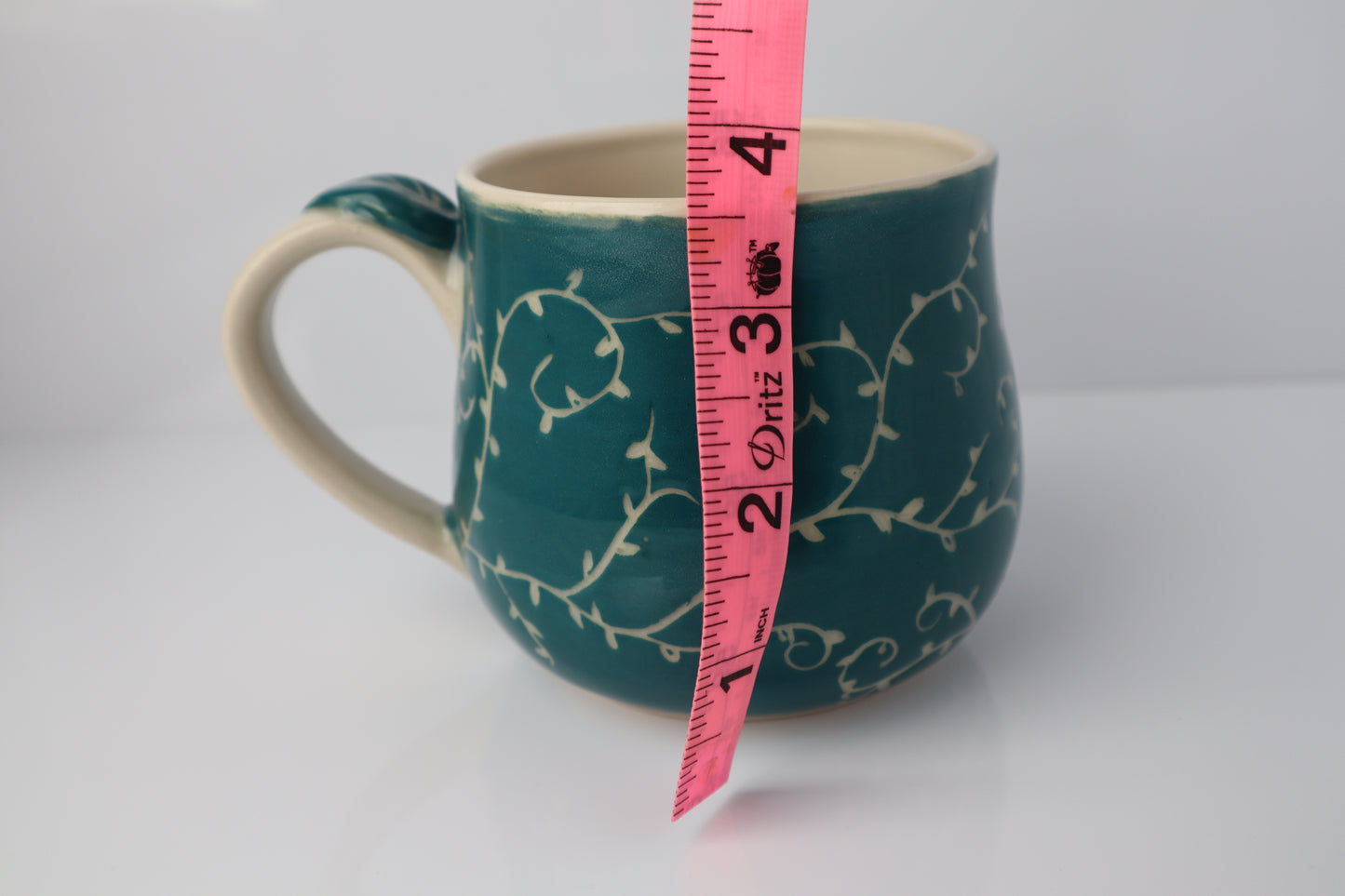 Teal mug with white floral pattern and pink measuring tape for scale on a white background