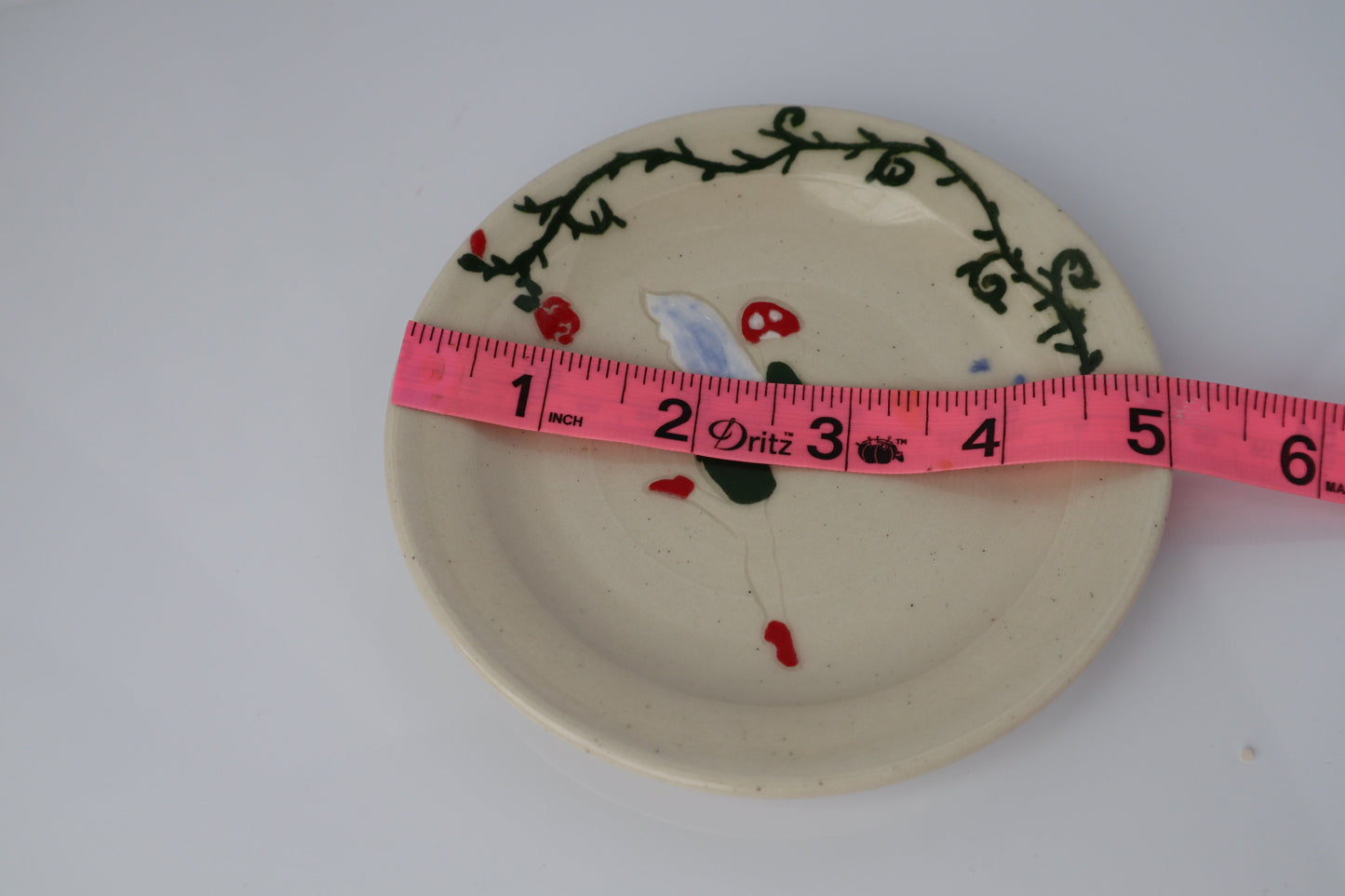 Small ceramic plate with floral design and a pink measuring tape for scale on a white background