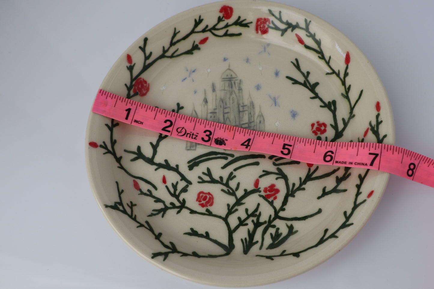 Decorative plate with floral and branch design, measured with a pink tape measure.
