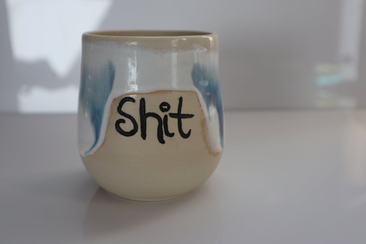 A handmade ceramic cup with a glossy finish, featuring a cream-colored base with blue brushstrokes and the word 'Shit' written in black marker across the front.