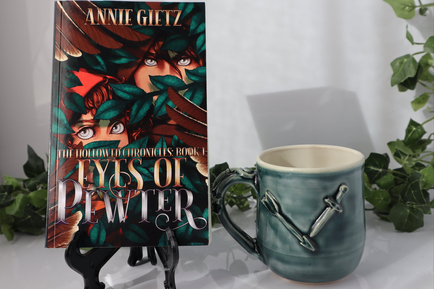 Book titled 'Eyes of Pewter' by Annie Gietz with a green mug with arrows and a sword on it, on a white surface.