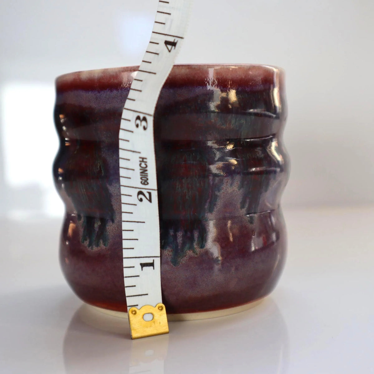 A small handmade ceramic cup with a pink and purple glaze, shown with a measuring tape vertically positioned inside the pot to indicate its height.