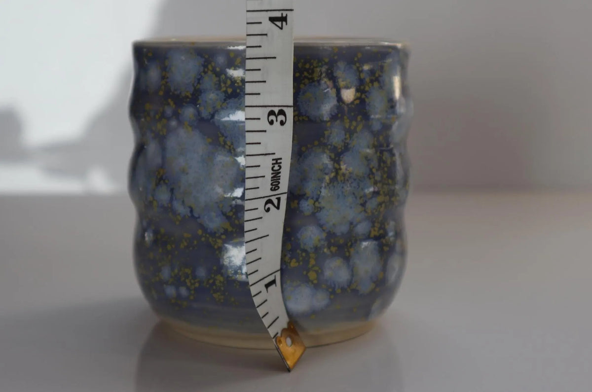 A small ceramic cup with a blue and white marbled glaze, shown with a measuring tape vertically aligned against it on a light gray background.