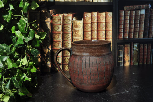 A  ceramic mug with a textured vertical pattern and a curved handle, set on a dark table in front of a bookshelf with antique books and green ivy.