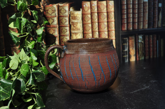 A handmade ceramic mug with a red clay left raw and blue crackle lines, set on a dark table in front of a bookshelf with books and green ivy.
