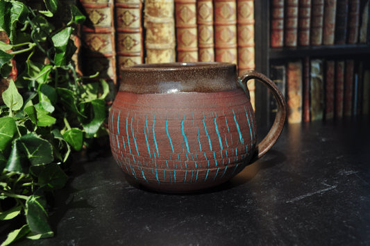 A handmade ceramic mug with a red clay body, decorated with vertical turquoise blue lines, set on a dark tabletop with a background of bookshelves and green foliage.