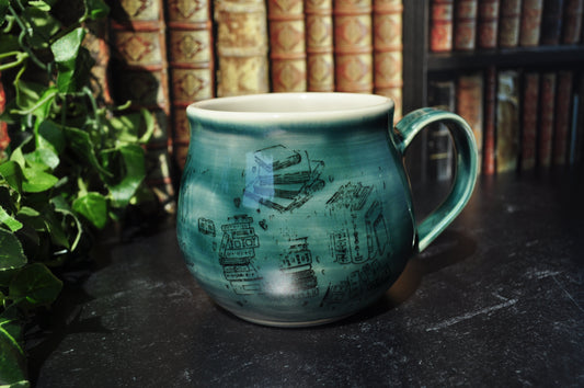 A handmade ceramic mug with a green glaze, decorated with illustrations of books. The mug is placed on a dark tabletop with a blurred background of bookshelves and green foliage.