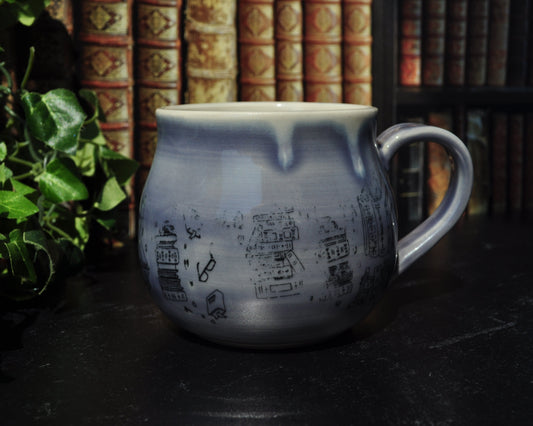 A purple ceramic mug with book designs the surface. Sitting on a black table top with a bookshelf in the background and green plants.