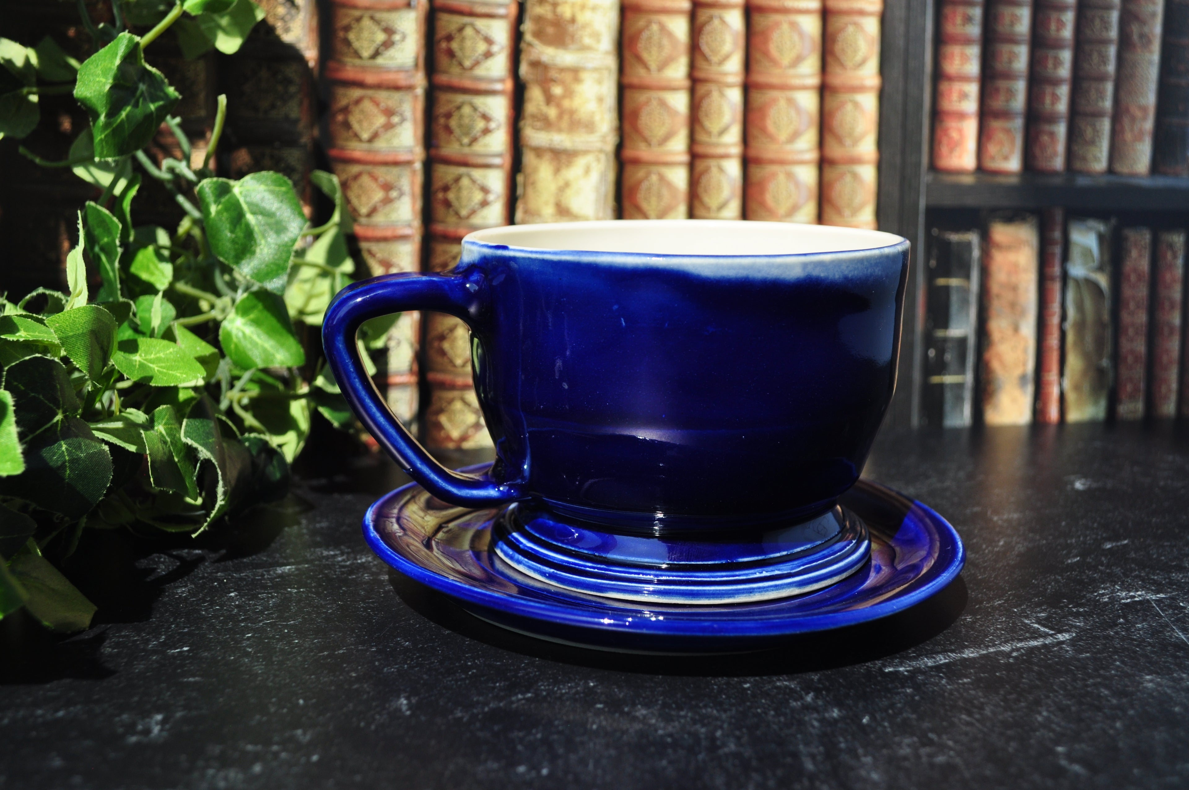 A glossy cobalt blue ceramic teacup and saucer set, styled on a dark tabletop with a background of vintage book spines and green foliage.