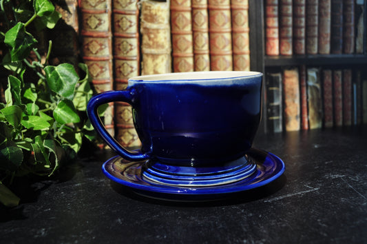 A glossy cobalt blue ceramic teacup and saucer set placed on a dark tabletop, with a blurred background of bookshelves and green leaves at the left edge.