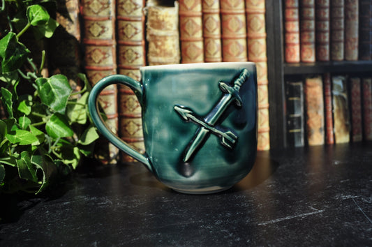 A green ceramic mug with a sculpted crossed-swords design on the side, with a blurred background of bookshelves and green foliage.