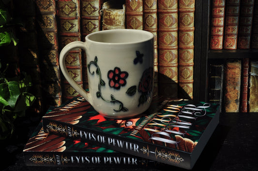 A white ceramic mug with floral vine and flower accents, resting on a stack of books in front of a dark bookshelf.