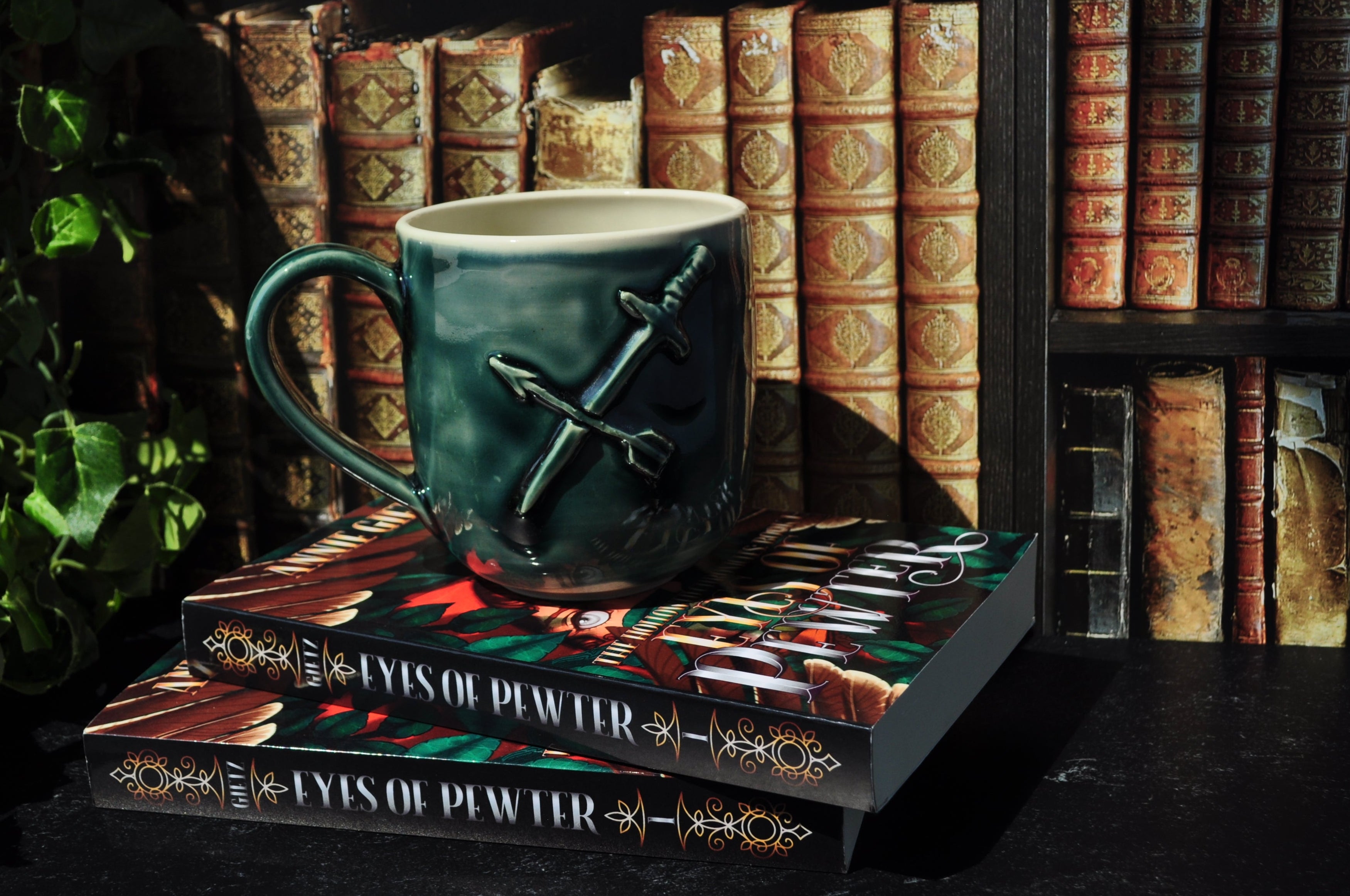 A green ceramic mug with a sword and arrow sculpted on the surface, resting on a stack of books in front of a bookshelf.