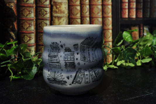 A small ceramic cup with  light purple and white and book imagery, set on a dark table with a background of bookshelves and green ivy.