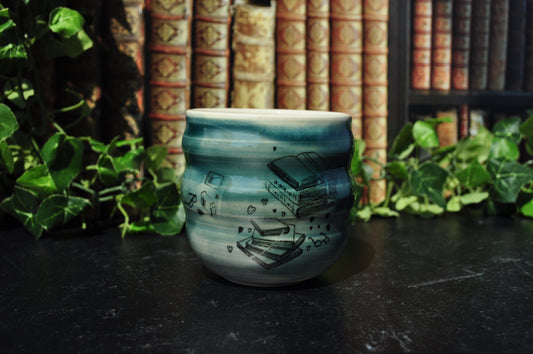 A small ceramic cup with green and white glaze and black illustrations of books, set on a dark table with books and green plants in the background.
