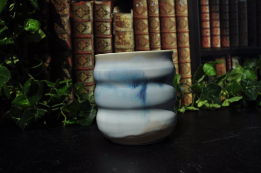 A ceramic cup with a wavy, ribbed design, featuring a white glaze with blue set against a background of vintage books and green ivy.