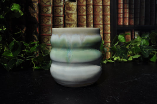 A small ceramic cup with a ribbed design, featuring a glossy glaze with white and pale green. It is set on a dark surface with a background of vintage books and green ivy.