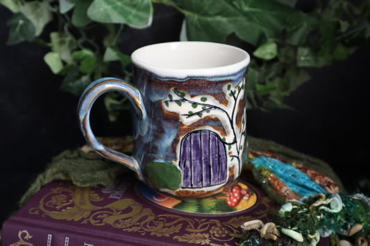 Ceramic mug shaped like a fairy house with a purple door, placed on a decorative surface with greenery in the background.