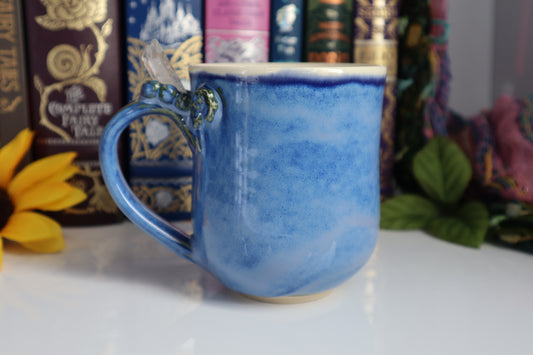Blue ceramic mug with a decorative handle on a white surface with books and plants in the background