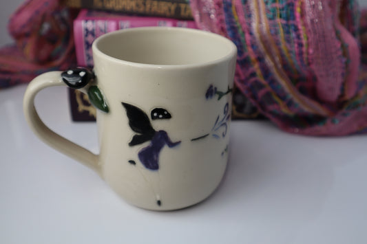 Beige mug with dark fairy design and a mushroom handle on a white surface