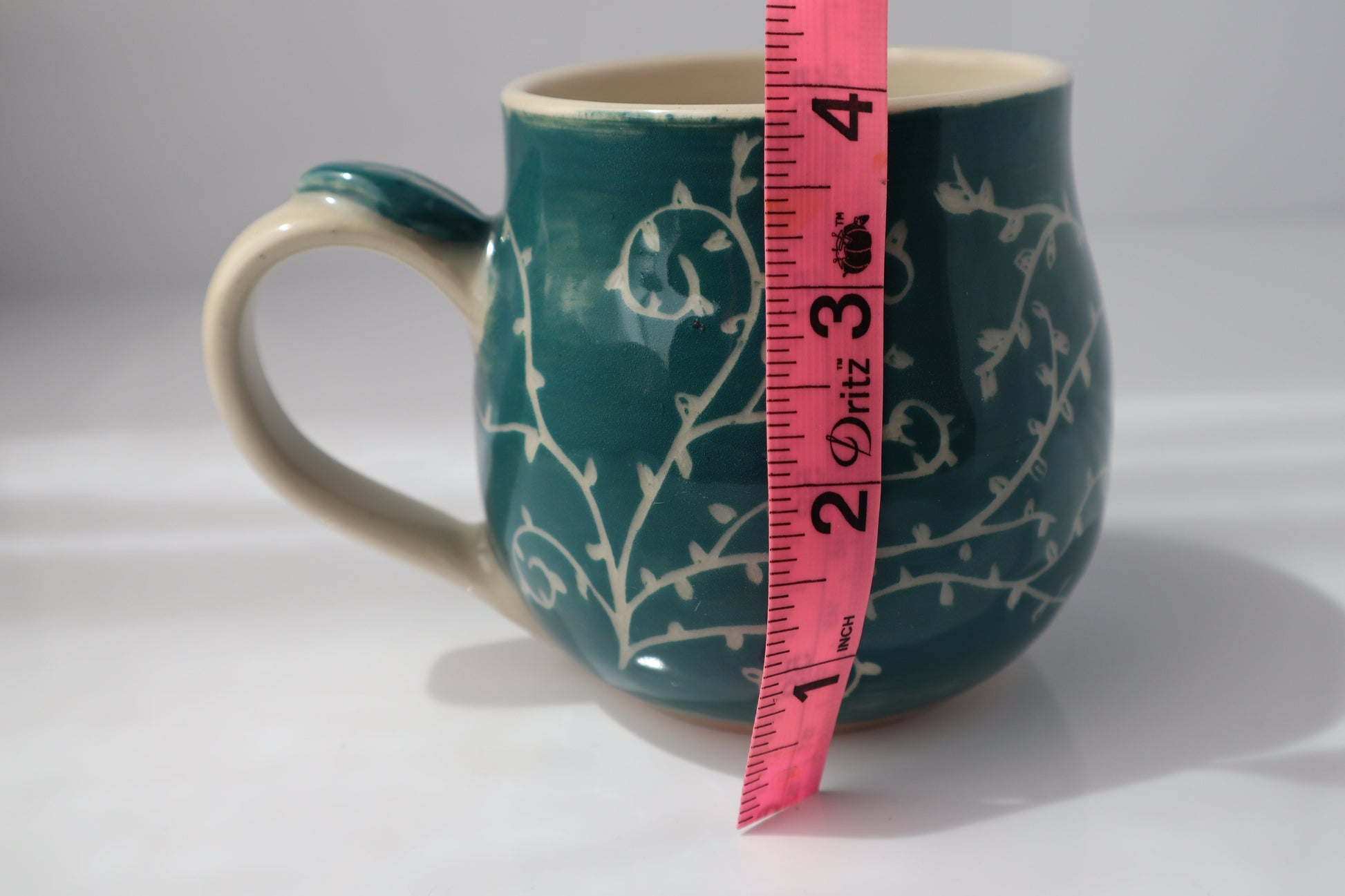 Teal mug with white floral pattern and pink measuring tape showing dimensions on a white background