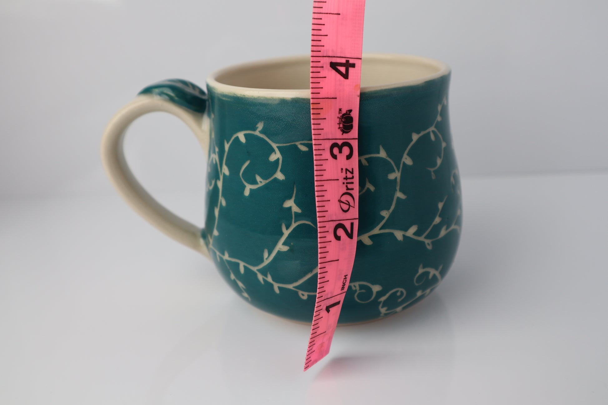 Teal mug with white floral pattern and pink measuring tape for scale on a white background