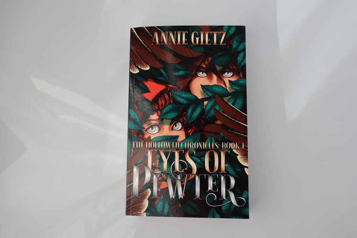 The book cover of  Eyes of Pewter by Annie Gietz, featuring a dark fantasy illustration of two characters with eyes hidden by green leaves, displayed on a light gray background.