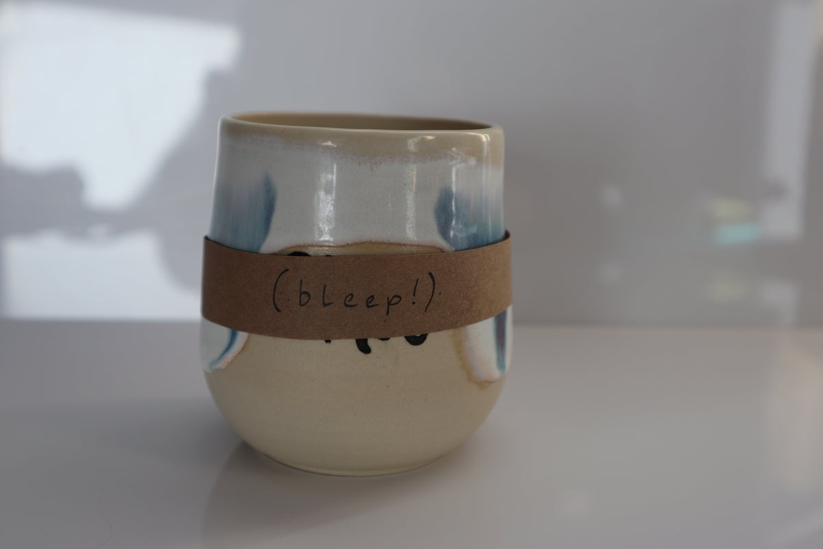A ceramic cup with a glossy cream and blue glazed finish, wrapped in a brown paper band that reads '(bleep!)' in black script, set against a softly blurred light gray background.