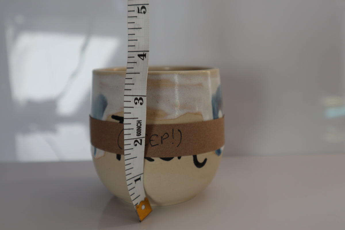 A small ceramic cup  with a beige base, and a brown paper band wrapped around the middle, shown with a measuring tape standing vertically to indicate size.