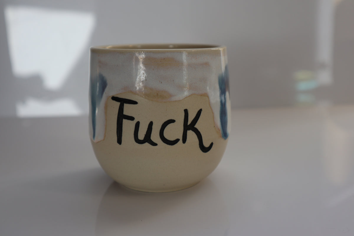 A handmade ceramic cup with a cream-colored glaze, blue painted accents, and the word 'Fuck' written in black marker on the front, set on a white surface.