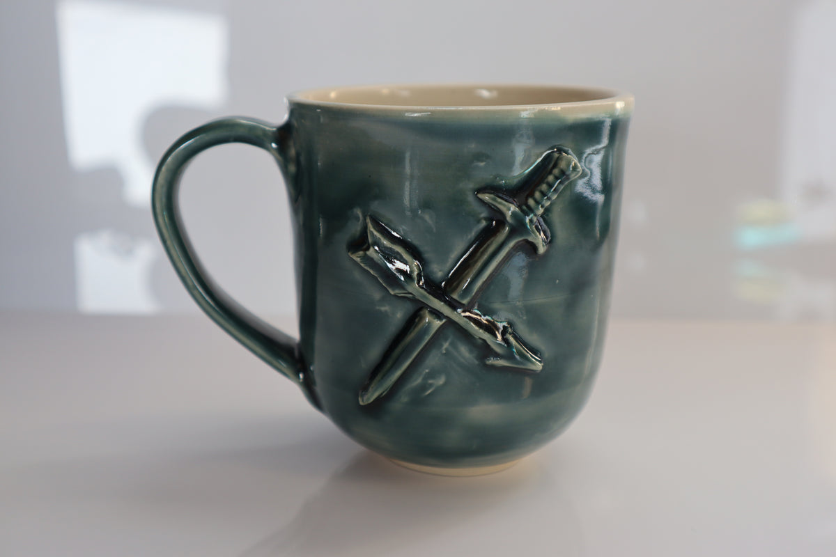 A handmade ceramic mug with a green glaze, featuring a raised relief of crossed sword and arrow on the front. The mug has a cream-colored interior and base, and is photographed on a white surface with a softly blurred background.