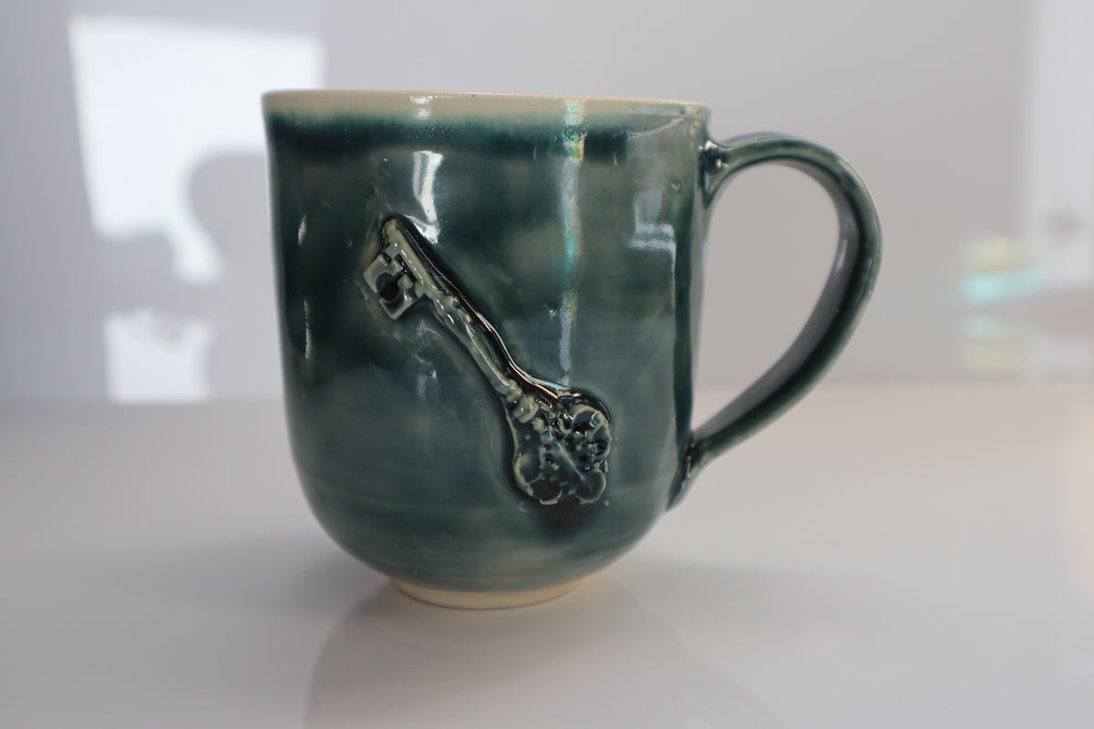 A handmade ceramic mug with a green glaze and a raised key design on the side, set against a softly blurred light gray background.