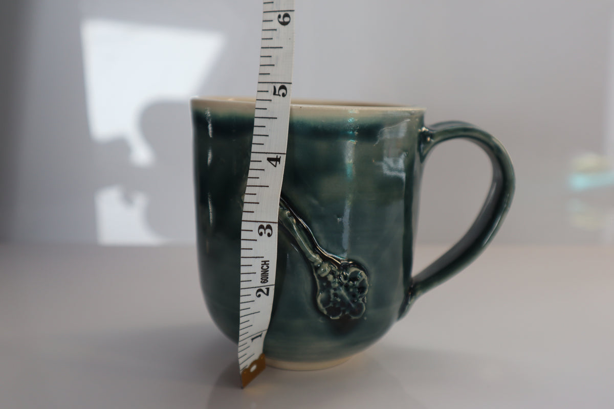 A handmade ceramic mug with a green glaze, shown next to a white measuring tape to indicate the scale of mug.