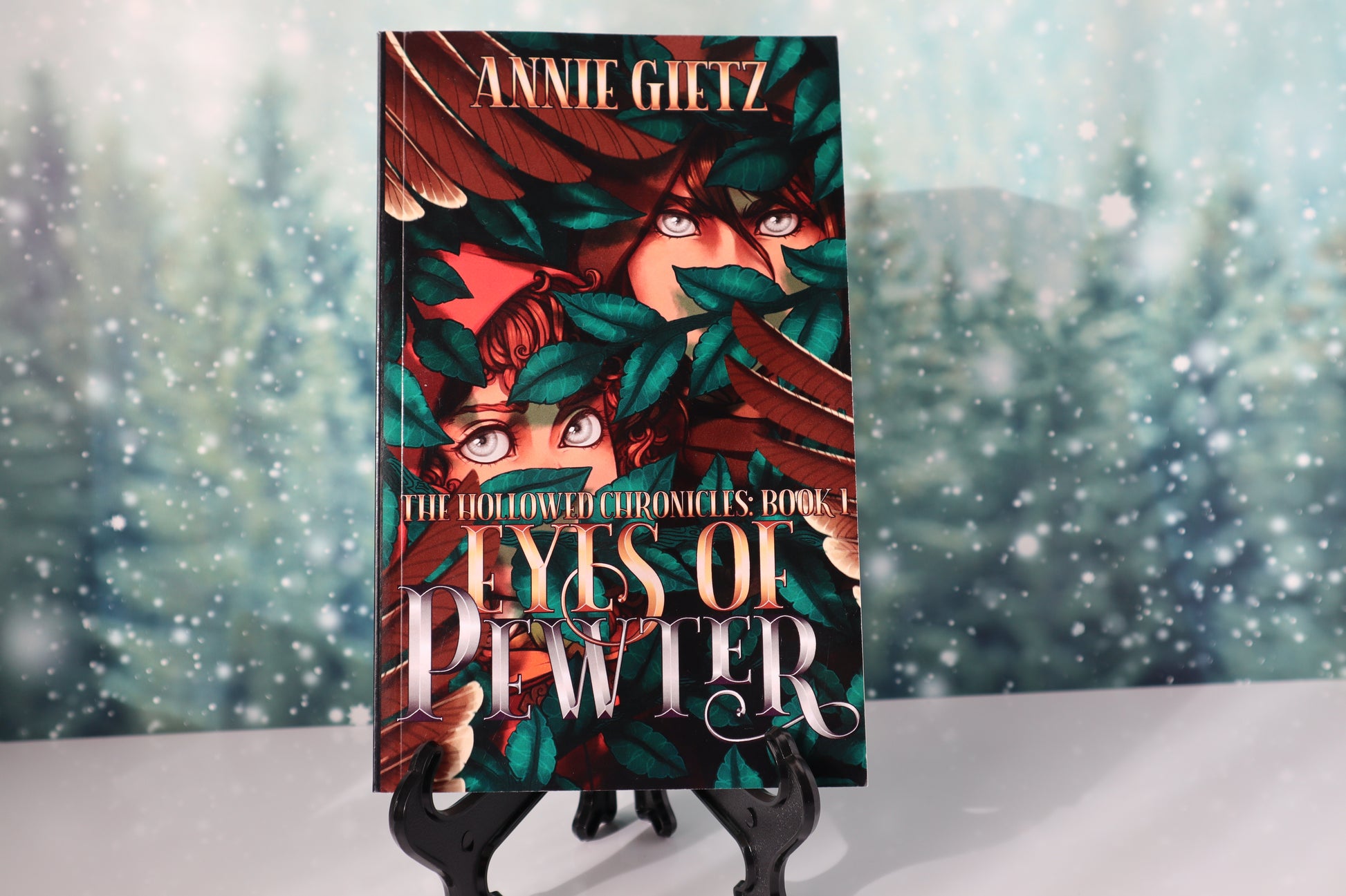 Book titled 'Eyes of Pewter' by Annie Gietz with a fantasy cover design of two hooded characters with silver eyes, on an book stand against a snowy background.