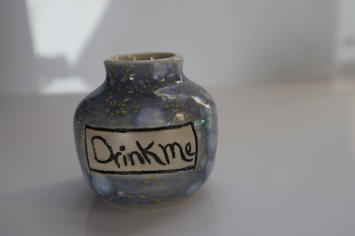 A small handmade ceramic bottle with a blue and gold speckled glaze, featuring a hand-painted label that reads 'Drink Me' in black underglaze, set against a neutral gray background.