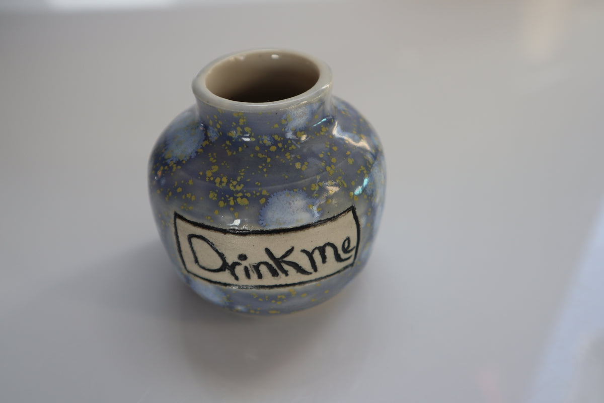 A small handmade ceramic bottle with a rounded body, a narrow neck, and a hand-painted cream label with the text 'Drink Me' written in black script.