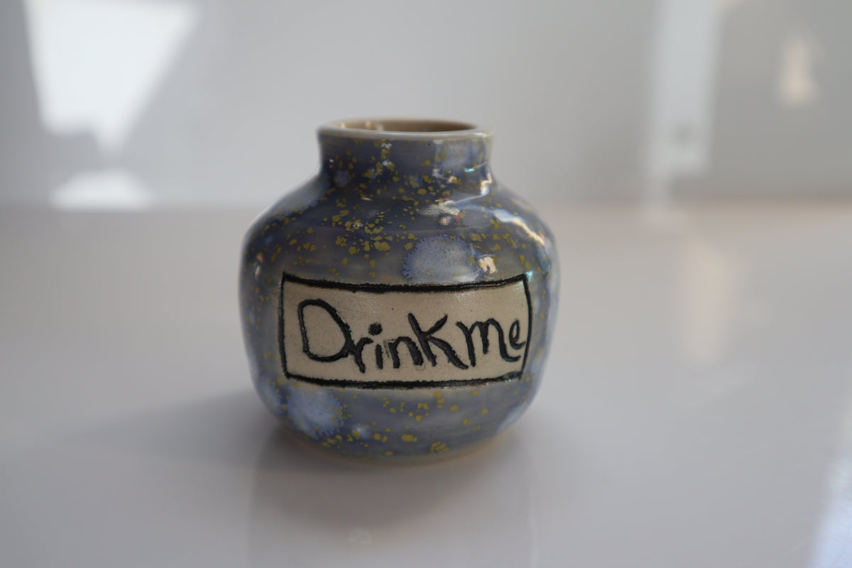A small handmade ceramic bottle with a lavender, light blue and yellow  glaze, featuring a hand-written label that reads 'Drink me' in black underglaze, set against a bright, softly blurred background.