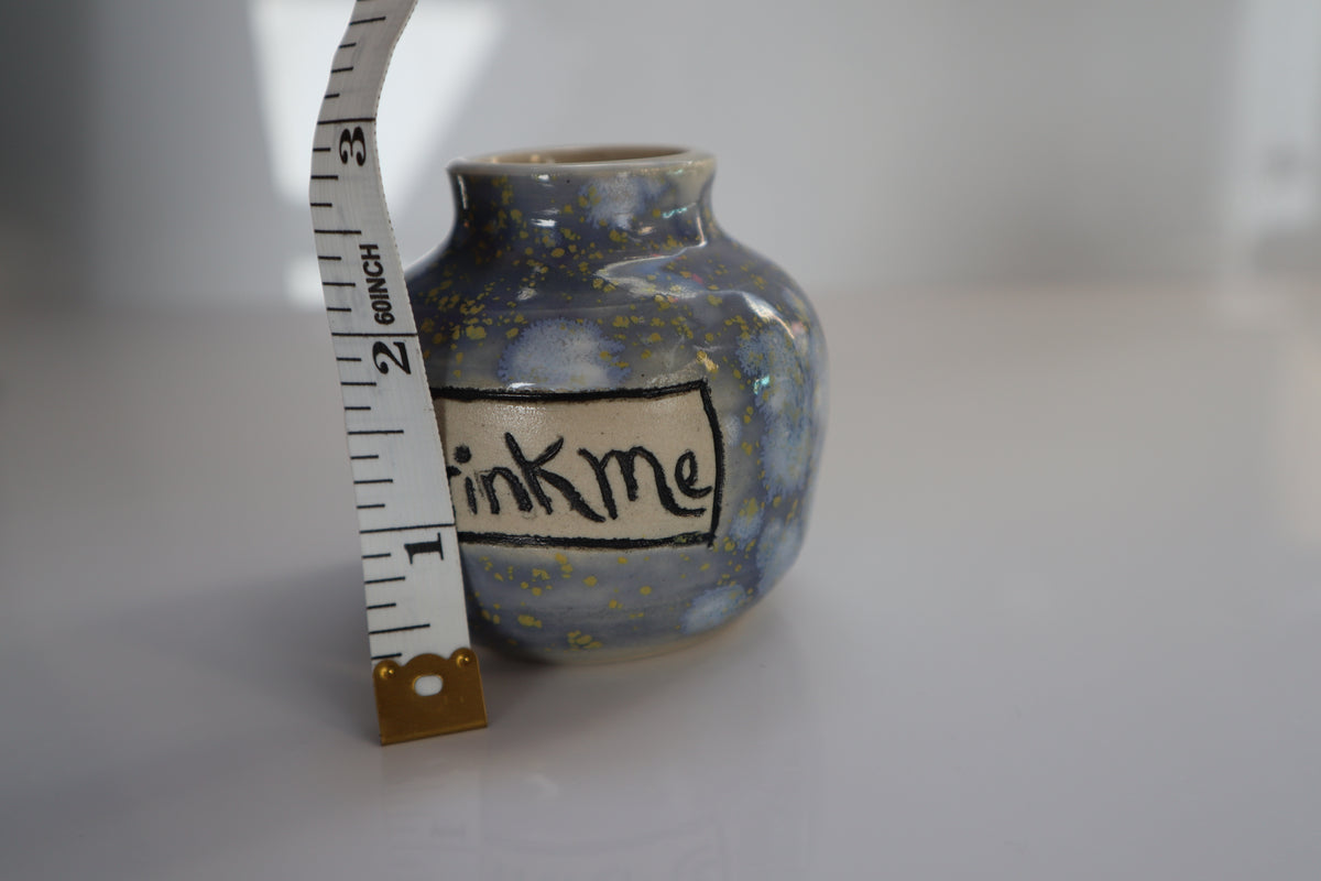 A small handmade ceramic pot with a blue and yellow speckled glaze, featuring a hand-painted label that reads 'Drink me' in black script. A measuring tape is positioned next to the pot to show its height against a neutral white background.