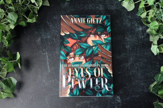 Book titled 'Eyes of Pewter' by Annie Gietz  with a fantasy cover of two hooded characters with silver eyes on a dark surface with green leaves around.