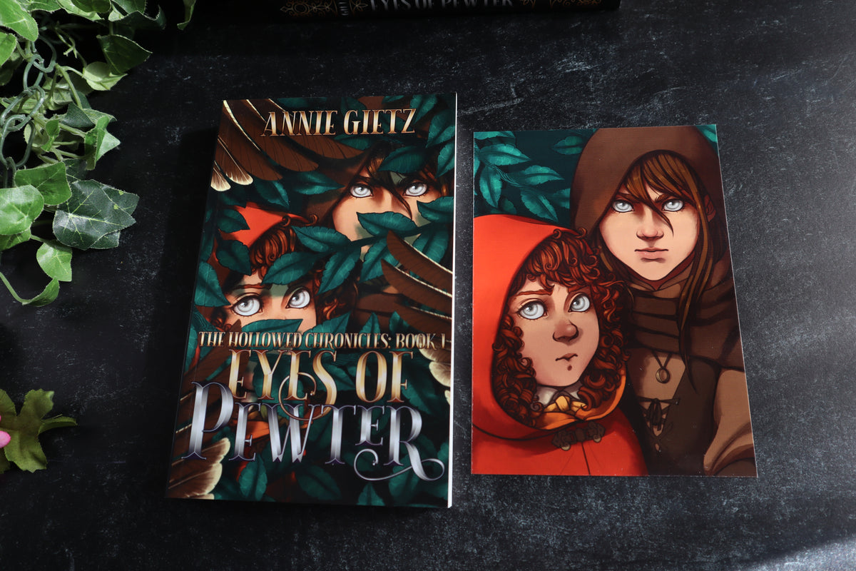 A book and a matching art print or card featuring illustrated characters from the book displayed side by side on a dark textured surface with decorative green leaves at the edge.