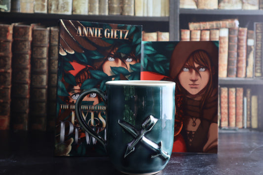 A dark teal ceramic mug with a sword and arrow design on the front, styled in front of a book and a printed character art featuring Kalahn and Piper, set against a blurred background of vintage bookshelves.