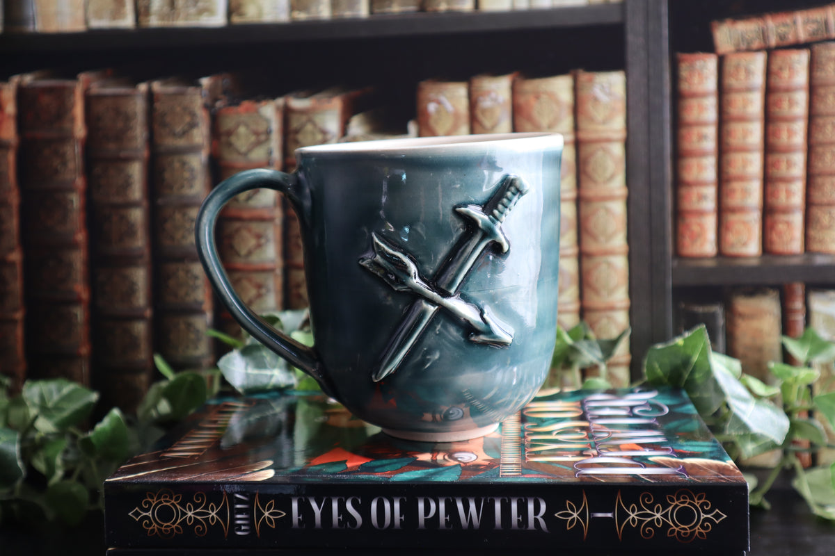 A green ceramic mug with a raised design of crossed sword and an arrow, displayed on a book in front of a blurred background of bookshelves.