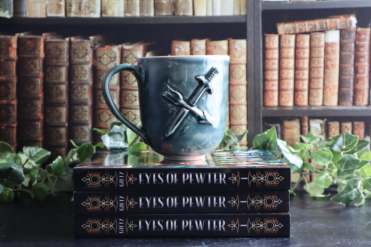 A green ceramic mug with crossed-sword and arrow design, displayed on top of two black 'Eyes of Pewter' books with green ivy leaves in the foreground, set against a background of bookshelves.