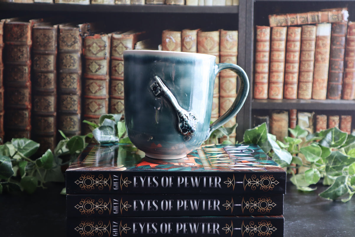 A green mug with a key on top of three stacked books titled 'Eyes of Pewter'. The background features a blurred library shelf filled with vintage books, with green ivy leaves framing the scene.