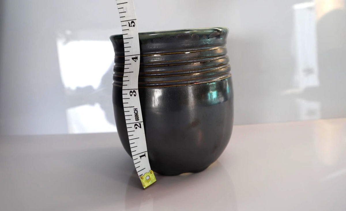 A small pewter glazed, ceramic vase with a glossy finish and horizontal ridges near the rim, shown on a light surface with a measuring tape standing alongside it for scale.