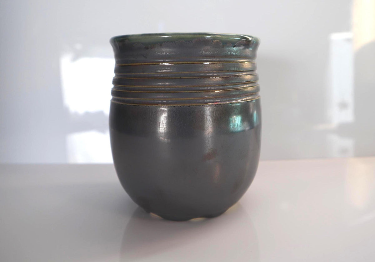 A small  pewter glazed, ceramic vase with a rounded body, a flared rim, and horizontal ridges around the upper half, shown on a light reflective surface against a white background.
