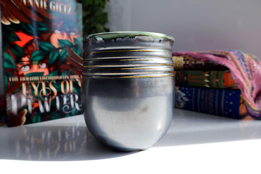 A metallic silver ceramic vase with a green glazed rim sits on a white surface, styled with books in the background.